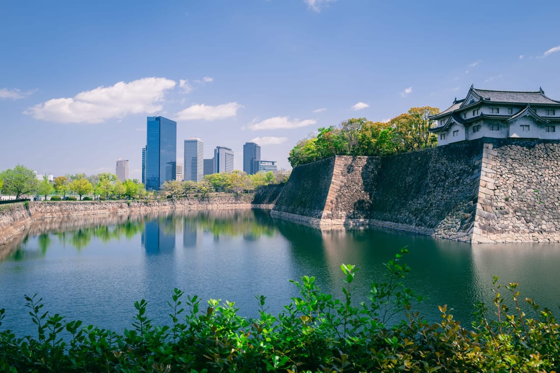 Picture taken in early April 2023 of Osaka Castle, surrounded by a moat, shrubbery, and skyscrapers off in the distance.